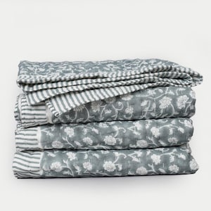 May include: A stack of folded quilts in shades of gray and white. The quilts feature floral and striped patterns. The top quilt is folded to show the striped pattern. The other quilts have a floral design.