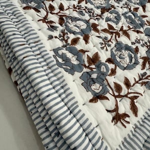May include: A folded white quilt with a floral pattern in shades of blue and brown. The quilt's edge features a striped pattern in white and blue. The quilt is made of a soft, quilted material.