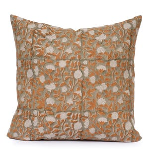 May include: Square decorative pillow with a floral pattern. The pillow features a light green background with a repeating design of white flowers and orange-brown leaves and stems. The pillow has a textured appearance.