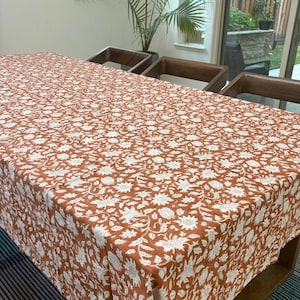 Puede incluir: Un mantel rectangular con un patrón floral blanco sobre un fondo terracota. El mantel está colocado sobre una mesa de madera, mostrando el intrincado diseño y el contraste de colores.