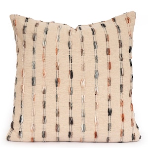 May include: A square throw pillow in a light beige fabric featuring vertical stripes of textured, raised yarn in shades of charcoal gray, soft cream, and muted terracotta. The woven material has a visible, consistent grid-like weave pattern.