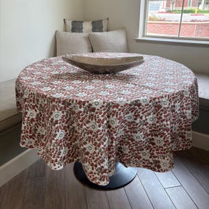 May include: A round table covered with a floral patterned tablecloth. The tablecloth features a repeating pattern of white flowers and red leaves on a white background. A wooden bowl sits in the center of the table. The table is approximately 48 inches in diameter.