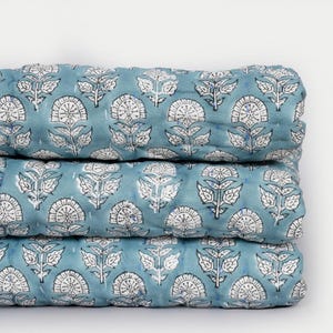 May include: A stack of three folded, teal-colored quilts. Each quilt features a repeating pattern of white floral designs with black outlines. The quilts are neatly stacked, showcasing the detailed print and soft texture of the fabric.