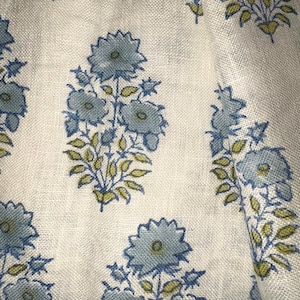 May include: Close-up of a white fabric with a repeating floral pattern. The design features clusters of blue flowers with yellow-green leaves. The fabric has a slightly textured appearance.