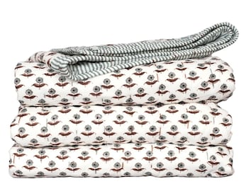 May include: A folded quilt featuring a white base with a repeating pattern of small dark gray flowers and brown stems. The top layer is partially folded back to reveal a reverse side with a pattern of thin, alternating dark gray and white stripes.