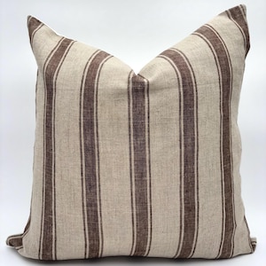 May include: A square pillow with a beige background and vertical brown stripes. The pillow has a soft, textured appearance, suggesting a linen or similar fabric. The stripes are evenly spaced and run the length of the pillow.