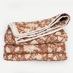 May include: A stack of three folded quilts with a floral pattern in shades of brown and cream. The quilts have a white border and are neatly stacked on a white background. The floral design is visible on each quilt.