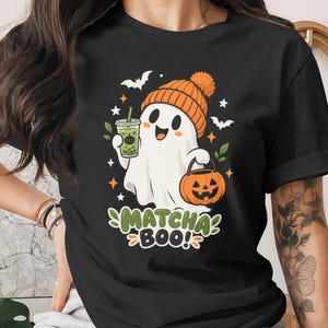 May include: A black short-sleeve t-shirt featuring a graphic of a white ghost wearing an orange beanie, holding a green matcha drink and an orange jack-o'-lantern bucket. The text "MATCHA BOO!" is printed below the ghost with small bats and stars.