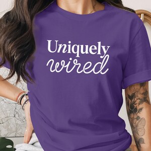 Könnte beinhalten: Ein violettes Kurzarm-T-Shirt mit dem weißen Schriftzug Uniquely wired auf der Vorderseite. Das Shirt hat einen Rundhalsausschnitt und umgeschlagene Ärmel, wobei der Text in einer Kombination aus Serifenschrift und Schreibschrift gedruckt ist.
