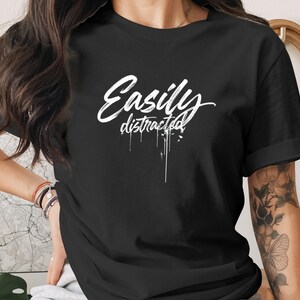 Könnte beinhalten: Ein schwarzes T-Shirt mit kurzen Ärmeln und dem weißen Schriftzug "Easily distracted" auf der Brust. Das Wort "Easily" ist in einer großen, stilisierten Schreibschrift gehalten, während "distracted" darunter mit einem tropfenden Farbeffekt dargestellt ist.