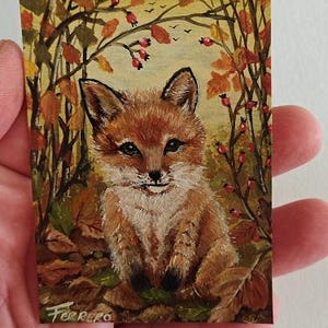 Può includere: Un piccolo dipinto rettangolare raffigurante una giovane volpe seduta tra il fogliame autunnale. La volpe ha il pelo arancione e bianco con orecchie e zampe dalle punte nere. Lo sfondo mostra alberi con foglie arancioni, bacche rosse e un cielo chiaro. Firmato Ferrero.