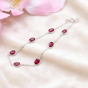 May include: A delicate silver bracelet featuring oval-cut, deep red gemstones. The gemstones are evenly spaced along a fine chain, creating a simple yet elegant design. The bracelet is displayed on a soft, white fabric, with pink flowers in the background.