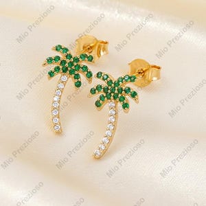 Könnte beinhalten: Goldfarbene Palmenohrringe mit grünen und weißen Edelsteinen. Die Palmwedel bestehen aus grünen Edelsteinen, und die Stämme sind mit weißen Edelsteinen besetzt. Die Ohrringe sind vor einem cremefarbenen Hintergrund platziert.