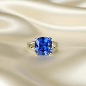 Peut inclure: Une bague en argent ornée d'une grosse pierre précieuse bleue vive de taille carrée. La pierre précieuse est le point central, sertie dans un sertissage classique à quatre griffes. La bague est présentée sur un tissu crème.