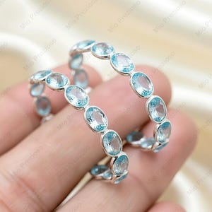 May include: Two silver rings, each set with oval, light blue gemstones. The rings are shown on a hand, highlighting the sparkling gems. The rings are a delicate piece of jewellery.