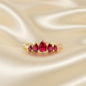 May include: A gold ring featuring a series of teardrop-shaped, vibrant red gemstones. The gemstones are arranged in a crown-like pattern, with the largest stone at the center. The ring is set against a soft, golden fabric.