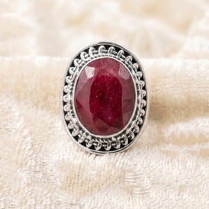 Könnte beinhalten: Ovaler Ring mit tiefrotem Rubin in silberfarbener, verzierter Fassung. Der facettierte Rubin ist von einem detaillierten, strukturierten Design umgeben.