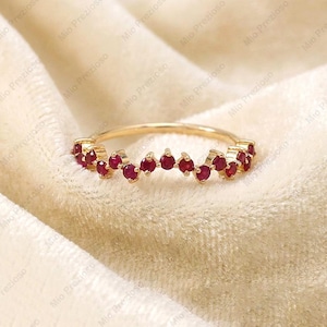Può includere: Un anello d'oro con una fila di piccole pietre preziose rosse e rotonde. Le pietre sono disposte a zig-zag lungo la parte superiore dell'anello. L'anello è presentato su un morbido tessuto color crema.