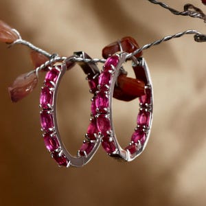 Könnte beinhalten: Ein Paar silberner Creolen, jede mit einer Reihe lebendiger, oval geschliffener, rosa Edelsteine besetzt. Die Ohrringe hängen an einem Draht mit rotbraunen und rosa Dekorationselementen vor einem warmen, neutralen Hintergrund.