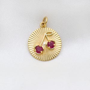 Op de afbeelding: Een goudkleurige hanger met een kersendesign. De hanger heeft een cirkelvormige vorm met een gestructureerd, zonnesterachtig patroon. Twee rode edelstenen stellen de kersen voor en er zit een kleine lus aan de bovenkant voor een ketting.