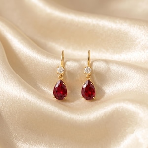 Puede incluir: Un par de pendientes colgantes de oro con una pequeña piedra redonda transparente en la parte superior y una piedra preciosa de color rojo intenso en forma de pera suspendida debajo. Tienen un cierre de gancho y un engaste de oro pulido.