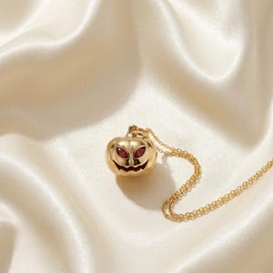May include: A gold-toned metal necklace featuring a pumpkin-shaped pendant. The pendant has a smooth, polished finish with cut-out details for the eyes and mouth, which are inlaid with deep red, almond-shaped stones, attached to a delicate chain.