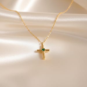 Pode incluir: Um colar dourado com um pingente de cruz. A cruz apresenta uma pequena pedra preciosa quadrada verde esmeralda no centro. A corrente do colar é delicada e o fundo é um tecido branco macio.