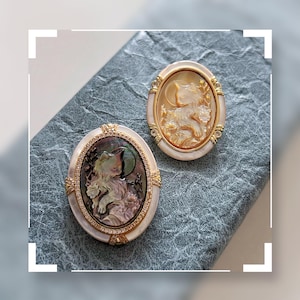Könnte beinhalten: Zwei ovale Katzen-Cameo-Broschen. Eine zeigt eine Katze mit Mond- und Blumenakzenten, eingefasst in einen goldfarbenen Rahmen mit Perlmutt-Rand. Die andere hat ein Katzendesign in einem goldfarbenen Rahmen mit weißem Rand.