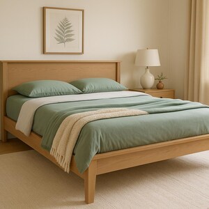 May include: A light wood bed frame with a sage green comforter and pillows. A cream-colored throw blanket is draped across the bed. A framed fern print hangs above the bed, and a potted plant sits nearby.