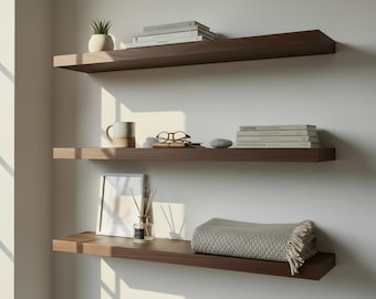 May include: Three dark brown floating shelves on a white wall. The top shelf has a small potted plant and books. The middle shelf holds a mug, glasses, and books. The bottom shelf displays a framed picture and a folded blanket.