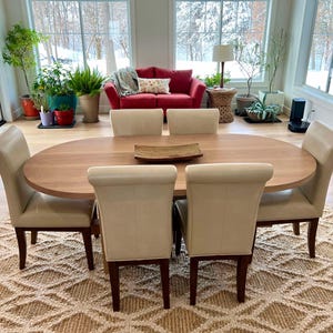 May include: A light wood oval dining table with six off-white chairs. The table is set with a wooden serving tray. The table is in a room with large windows and a red sofa.