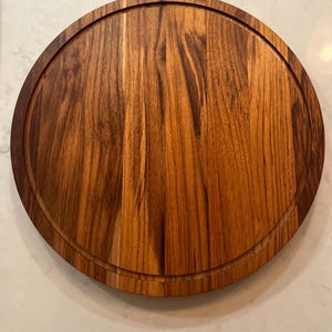 May include: A round wooden cutting board with a groove around the edge. The board is made of a light brown wood with darker brown grain patterns. The board is approximately 12 inches in diameter.