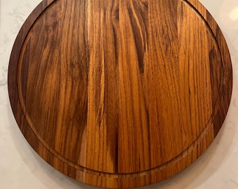 May include: A round wooden cutting board with a groove around the edge. The board is made of a light brown wood with darker brown grain patterns. The board is approximately 12 inches in diameter.