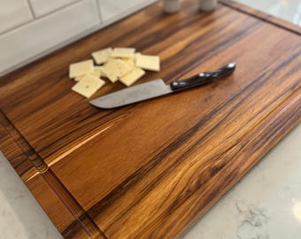 May include: A rectangular wooden cutting board with a juice groove, a knife, and cheese. The board is made of wood with a rich brown grain. Two mushroom-shaped salt and pepper shakers are in the background.