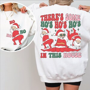 May include: White sweatshirt with a Christmas-themed graphic. The graphic features three Santa Clauses in various poses with the text "There's some Ho's Ho's Ho's in this house".