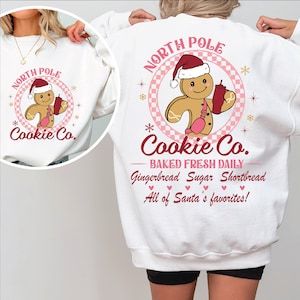 May include: White sweatshirt with a pink and red graphic of a gingerbread man holding a cup of hot chocolate. The graphic says "North Pole Cookie Co. Baked Fresh Daily Gingerbread Sugar Shortbread All of Santa's favorites!"
