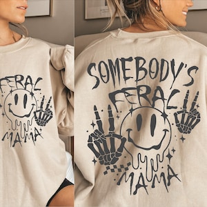 May include: A beige sweatshirt with a black graphic print of a smiley face with dripping eyes and the text "Somebody's Feral Mama" with two skeleton hands on either side of the smiley face.