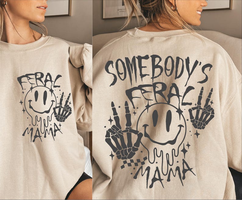 May include: A beige sweatshirt with a black graphic print of a smiley face with dripping eyes and the text "Somebody's Feral Mama" with two skeleton hands on either side of the smiley face.