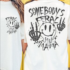 May include: White t-shirt with a black graphic print of a smiley face with dripping eyes and the text "Somebody's Feral Mama" on the back. The graphic also includes two skeleton hands with peace signs.