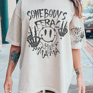 May include: A light beige oversized t-shirt with a black graphic print of a smiley face with dripping eyes, skeleton hands, and the text "Somebody's Feraci Mama".