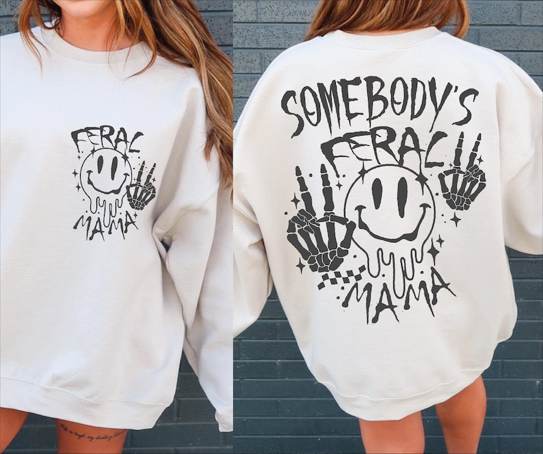 May include: White sweatshirt with black graphic print. The graphic features a smiling face with the text "Somebody's Feral Mama" and skeletal hands.
