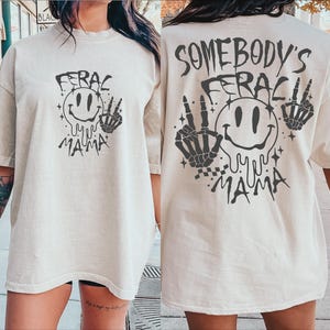 May include: Two oversized, light beige t-shirts with a black graphic print. The graphic features a smiling face with dripping eyes, two skeletal hands, and the text "Feral My Mama". One shirt has the text "Somebody's" above the graphic.