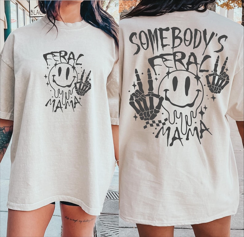May include: Two oversized, light beige t-shirts with a black graphic print. The graphic features a smiling face with dripping eyes, two skeletal hands, and the text "Feral My Mama". One shirt has the text "Somebody's" above the graphic.