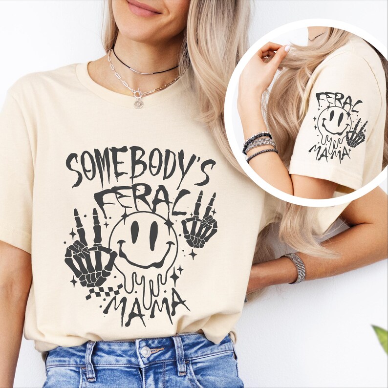 May include: A beige t-shirt with a black graphic design that says "Somebody's Feral Mama" with a smiley face and skeleton hands.