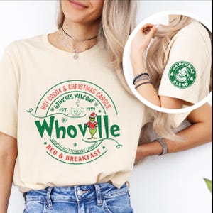 Peut inclure: Un t-shirt cr&egrave;me avec un motif vert et rouge qui dit "Hot Cocoa & Christmas Carols *Grinches Welcome* Est. 1954 Whoville *Located next to Mount Crumpit* Bed & Breakfast" avec un personnage de dessin anim&eacute; du Grinch. La manche droite a un cercle vert avec le texte "Grinchmas Blend" et un personnage de dessin anim&eacute; du Grinch.