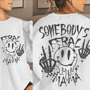 May include: White long-sleeve sweatshirt with a black graphic print of a smiley face with dripping eyes and the text "Somebody's Feraci Yu Yu Mama" with two skeleton hands making peace signs.