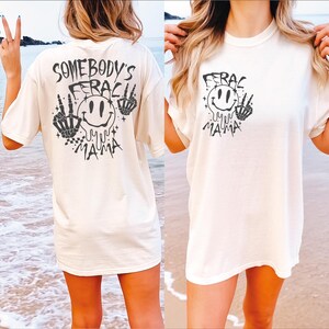 May include: White oversized t-shirt with a black graphic print of a smiley face with the text "Somebody's Ferac Mama" on the back and "Ferac Mama" on the front.