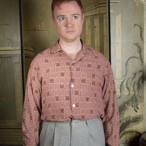 Puede incluir: Camisa de manga larga color melocotón con un estampado geométrico en negro y morado. La camisa se combina con pantalones grises. El conjunto es de estilo vintage.