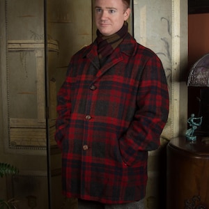 Könnte beinhalten: Ein rot-schwarz karierter Wollmantel mit Knopfleiste und Taschen. Der Mantel wird über einem braunen und beigefarbenen Schal getragen. Der Mantel ist ein klassischer Stil, geeignet für kaltes Wetter.