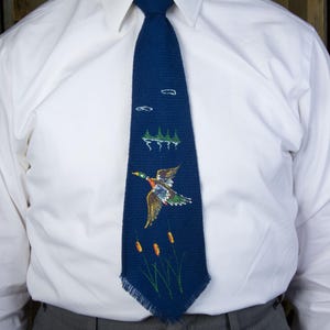 May include: A navy blue tie featuring a detailed embroidered design of a flying duck, cattails, and a small landscape. The tie is worn over a white dress shirt and gray trousers. The tie's design includes shades of green, brown, and yellow.